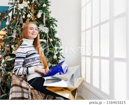 Happy girl with packages after christmas shopping. Smiling young woman in casual at home interior with decorated xmas tree and gift boxes posing on camera. Winter holidays sales. Copy spase 138403825