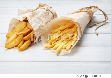 Potato wedges and french fries wrapped in brown wrapping paper. Fast food take away at white shabby chic wood. Fried potatoes with tomato sauce. Chips, potato slices. Top view, flat lay, closeup. Potato wedges and french fries wrapped in brown wrapping paper. Fast food take away at white shabby chic wood. Fried potatoes with tomato sauce. Chips, potato slices. Top view, flat lay, closeup. 138403862