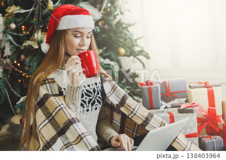 Girl with laptop sitting on the floor at decorated christmas tree background. Young woman drinking coffee and writing greeting emails online in holiday home atmosphere among present boxes. Copy space 138403964