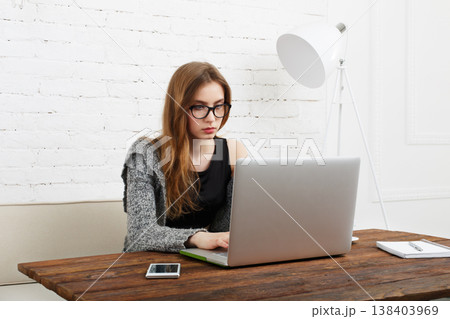 A young woman sits at a wooden table in a minimal room, focused on her laptop. She has long hair and wears glasses. A smartphone is next to her, and bright light fills the space. A young woman sits at a wooden table in a minimal room, focused on her laptop. She has long hair and wears glasses. A smartphone is next to her, and bright light fills the space. 138403969