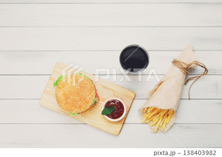 Burger, Chips and cola. Hamburger and french fries wrapped in brown wrapping paper. Fast food take away at white shabby chic wood. Hamburger with tomato sauce. Top view, flat lay with copyspace Burger, Chips and cola. Hamburger and french fries wrapped in brown wrapping paper. Fast food take away at white shabby chic wood. Hamburger with tomato sauce. Top view, flat lay with copyspace 138403982