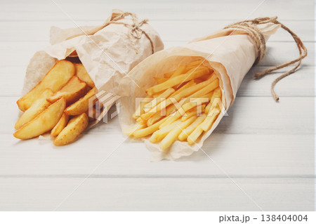 Potato wedges and french fries wrapped in brown wrapping paper. Fast food take away at white shabby chic wood. Fried potatoes with tomato sauce. Chips, potato slices. Top view, flat lay, closeup. Potato wedges and french fries wrapped in brown wrapping paper. Fast food take away at white shabby chic wood. Fried potatoes with tomato sauce. Chips, potato slices. Top view, flat lay, closeup. 138404004