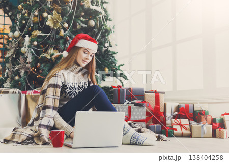 Girl with laptop sitting on the floor at decorated christmas tree background. Young woman drinking coffee and writing greeting emails online in holiday home atmosphere among present boxes. Copy space 138404258