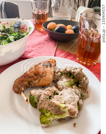 A plate features baked rabbit and broccoli with sauce. A fresh green salad sits nearby. Glasses of tea are filled. Apricots and plums are on the table for dessert 138404557