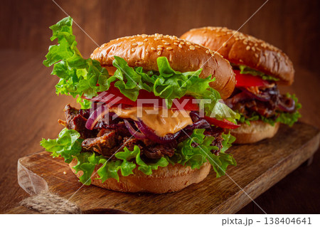 Two burgers with torn beef, tomatoes, fried onions and sauce, laid out on a wooden board, there are no people. Two burgers with torn beef, tomatoes, fried onions and sauce, laid out on a wooden board, there are no people. 138404641