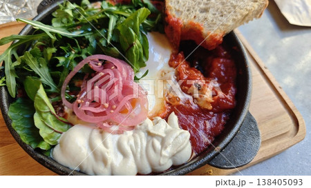 Baked eggs in rich tomato sauce with fresh greens, pickled onions, and a slice of bread presented in a cast iron skillet on a wooden surface Baked eggs in rich tomato sauce with fresh greens, pickled onions, and a slice of bread presented in a cast iron skillet on a wooden surface 138405093