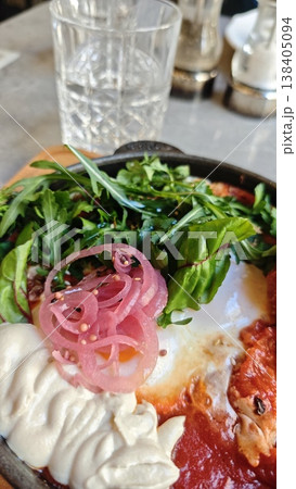 Dish of baked eggs served in a cast iron skillet with fresh greens, pickled onions, and a dollop of cream on a wooden board with a glass of water in the background 138405094