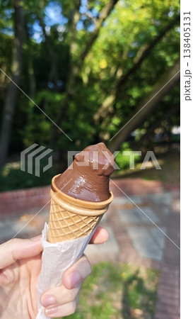 Hand holding a chocolate ice cream cone with a blurred park setting in the background, showcasing trees and a paved walkway under natural light Hand holding a chocolate ice cream cone with a blurred park setting in the background, showcasing trees and a paved walkway under natural light 138405131
