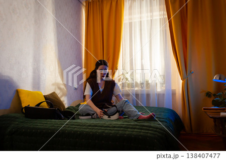 Focused female in VR goggles explores immersive content on bed in softly lit bedroom. Indoor gaming and digital life. Wide shot 138407477