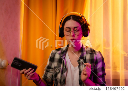 Portrait of happy teen listening to energetic music through headphones while relaxing in her bedroom. Evening glow, cozy mood and youthful lifestyle Portrait of happy teen listening to energetic music through headphones while relaxing in her bedroom. Evening glow, cozy mood and youthful lifestyle 138407492