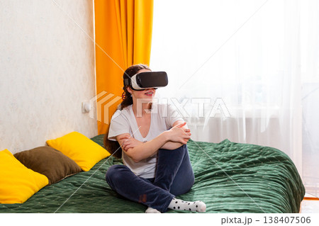 Female embracing the evolution of virtual reality gaming while relaxing indoors, testing high-end simulations, sitting on bed 138407506