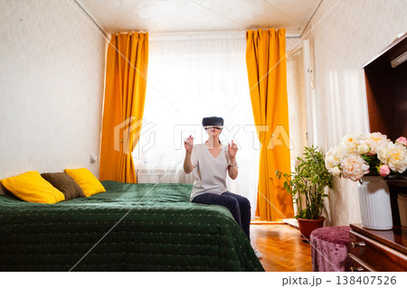 Woman wearing virtual reality headset while relaxing on bed in stylish bedroom, experiencing futuristic interactive media. Wide shot Woman wearing virtual reality headset while relaxing on bed in stylish bedroom, experiencing futuristic interactive media. Wide shot 138407526