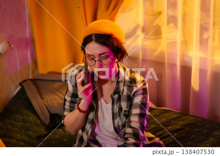 Casual female teen sitting on bed, engaged in phone call in sunset ambiance. Cozy home mood and private talk atmosphere. Mid shot 138407530