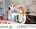 A happy Easter scene with a mother and daughter putting on paper bunny masks at the kitchen table while playfully preparing for the holiday together. 138407554