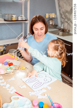 A Caucasian woman and her daughter craft holiday ornaments together, cutting decorations at the table and celebrating Easter traditions with children. Vertical 138407556