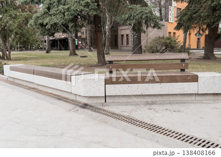 Modern Urban Park Bench With Concrete Base And Wooden Slats Amidst Green Trees And City Buildings In Overcast Weather 138408166