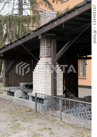 Rustic Outdoor Brick Chimney Structure With Dark Wooden Roof And Concrete Steps In Garden Setting Featuring Ornate Metal Fence Details And Mossy Trees Background 138408167