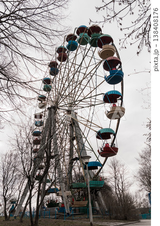 Vintage Colorful Ferris Wheel Amidst Bare Trees In Overcast Amusement Park Scene With Red Blue And Green Passenger Cars Standing Tall Against Gray Sky Vintage Colorful Ferris Wheel Amidst Bare Trees In Overcast Amusement Park Scene With Red Blue And Green Passenger Cars Standing Tall Against Gray Sky 138408176