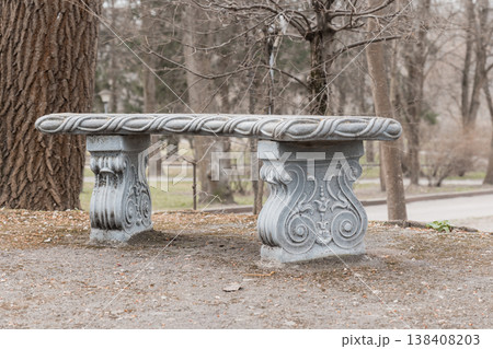 Ornate Gray Stone Bench With Intricate Carvings And Rope Detail In Quiet Park Surrounded By Bare Trees During Early Spring Seasonal Transition 138408203