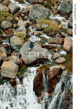 A photographer girl sits on a rock at the edge of the Battery waterfall in Teriberka, Arctic landscape with a view of a flowing waterfall in the northern fjord, Arctic, Russia, Murmansk region 138408242