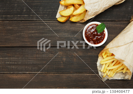 Potato wedges and french fries wrapped in brown wrapping paper. Fast food take away at rustic wood background. Choice of fried potatoes with tomato sauce. Chips, potato slices. Top view, copyspace Potato wedges and french fries wrapped in brown wrapping paper. Fast food take away at rustic wood background. Choice of fried potatoes with tomato sauce. Chips, potato slices. Top view, copyspace 138408477