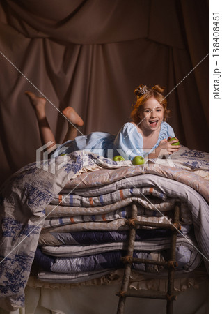 Little girl in blue dress lying on stacked mattresses with apples, princess fairy tale concept 138408481
