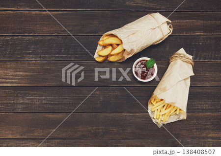 Potato wedges and french fries wrapped in brown wrapping paper. Fast food take away at rustic wood background. Choice of fried potatoes with tomato sauce. Chips, potato slices. Top view, copyspace Potato wedges and french fries wrapped in brown wrapping paper. Fast food take away at rustic wood background. Choice of fried potatoes with tomato sauce. Chips, potato slices. Top view, copyspace 138408507