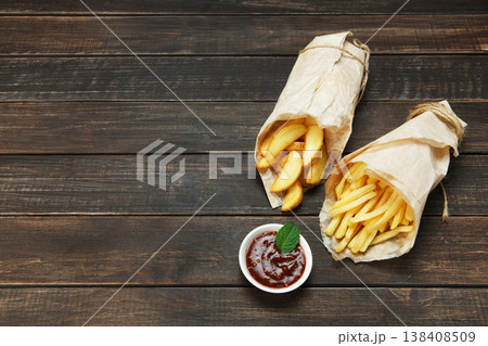 Potato wedges and french fries wrapped in brown wrapping paper. Fast food take away at rustic wood background. Choice of fried potatoes with tomato sauce. Chips, potato slices. Top view, copyspace 138408509