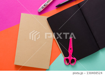 Stationery at modern creative work space. Stylish open notepad with black pages, pink scissors, office knife, marker and craft envelope on abstract multicolored background, top view, copy space Stationery at modern creative work space. Stylish open notepad with black pages, pink scissors, office knife, marker and craft envelope on abstract multicolored background, top view, copy space 138408524