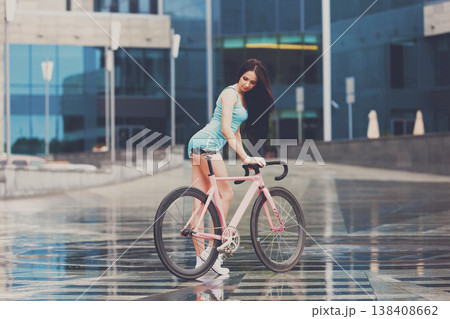 Sexy girl on bicycle. Young slim sexy sportive woman in blue shorts and white snickers long-haired, sensual posing on pink fix bicycle at grey background in urban city enviroment. Soft toned. Sexy girl on bicycle. Young slim sexy sportive woman in blue shorts and white snickers long-haired, sensual posing on pink fix bicycle at grey background in urban city enviroment. Soft toned. 138408662