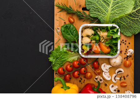 Healthy vegetarian food delivery background. Fresh organic vegetables on black. Natural food in foil box and wooden cutting board with copy space. Healthy vegetarian food delivery background. Fresh organic vegetables on black. Natural food in foil box and wooden cutting board with copy space. 138408796