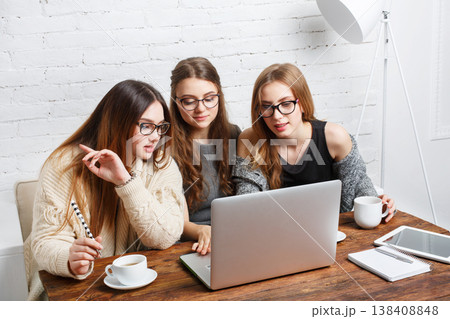 Three friends share ideas as they work together on a laptop. They sit at a wooden table with coffee cups, focused on the screen and engaged in discussion. Three friends share ideas as they work together on a laptop. They sit at a wooden table with coffee cups, focused on the screen and engaged in discussion. 138408848