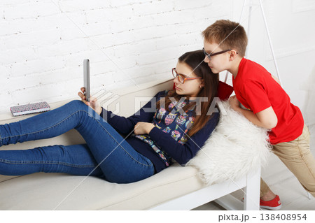 Two kids are sitting on a couch in a bright room. One child lounges on her back, looking at a tablet. The other child stands close, watching the screen with interest. Two kids are sitting on a couch in a bright room. One child lounges on her back, looking at a tablet. The other child stands close, watching the screen with interest. 138408954