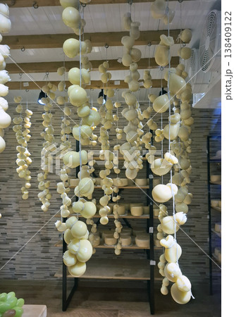 A variety of caciocavallo and scamorza cheeses hang from strings in a rustic cellar in Italy. The room has wooden beams and features a shelf with more cheeses. A variety of caciocavallo and scamorza cheeses hang from strings in a rustic cellar in Italy. The room has wooden beams and features a shelf with more cheeses. 138409122
