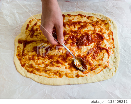 Top view of woman hands spreading organic tomato sauce on handmade pizza dough with spoon, traditional italian recipe preparation in cozy home kitchen, domestic culinary hobby lifestyle photography Top view of woman hands spreading organic tomato sauce on handmade pizza dough with spoon, traditional italian recipe preparation in cozy home kitchen, domestic culinary hobby lifestyle photography 138409211