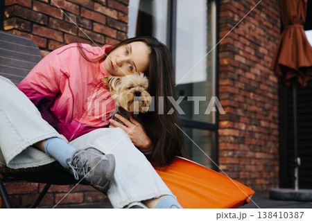 Young woman in pink windbreaker hugging yorkshire terrier outdoors, sitting on folding chair near brick house, authentic pet owner lifestyle, casual autumn outfit, bonding moment with dog, real life. Young woman in pink windbreaker hugging yorkshire terrier outdoors, sitting on folding chair near brick house, authentic pet owner lifestyle, casual autumn outfit, bonding moment with dog, real life. 138410387