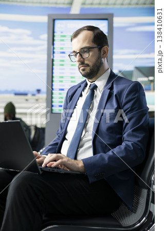 Man CEO in formal wear waiting in airport terminal lounge area, reviewing important documents on laptop before departure. International business and busy lifestyle of professional frequent flyer. 138410631