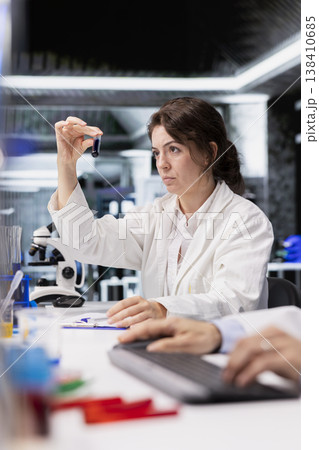Analytical chemist using laboratory glassware filled with indicator solution at workstation in research facility. Lab employee looking at cylinder with chemical fluid used for experiments 138410685