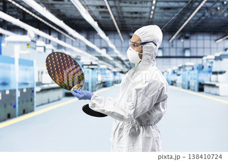 Man in bunny suit evaluating silicon wafer in brightly lit manufacturing plant cleanroom. Engineer checking semiconductor substrate nanometer circuit in modern foundry facility Man in bunny suit evaluating silicon wafer in brightly lit manufacturing plant cleanroom. Engineer checking semiconductor substrate nanometer circuit in modern foundry facility 138410724