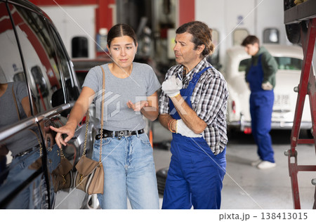 Worried girl points to scratches on car after accident to auto mechanic at auto repair shop Worried girl points to scratches on car after accident to auto mechanic at auto repair shop 138413015
