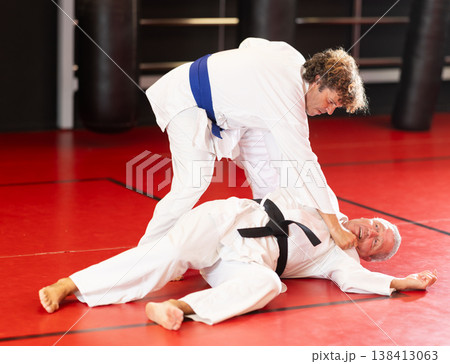 Concentrated blue-belt middle-aged aikido fighter in white gi honing fighting techniques during training kumite with older male partner in dojo 138413063