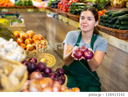 Female grocery store worker arranges red onion and other vegetables on counter and display case Female grocery store worker arranges red onion and other vegetables on counter and display case 138413142