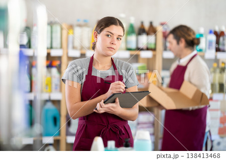 Girl seller, with tablet in hands, make inventory in store. Man employee in background Girl seller, with tablet in hands, make inventory in store. Man employee in background 138413468