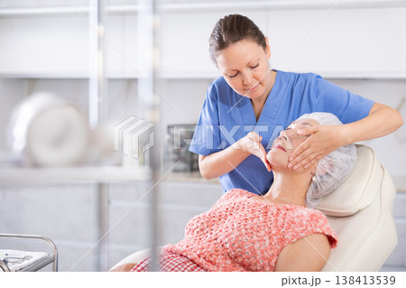 Elderly woman lies in chair during appointment with cosmetologist and enjoys facial massage 138413539