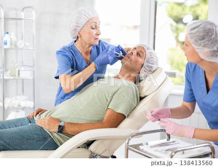 Elderly woman doctor gives injection of botulinum toxin in lip area to man, female nurse assist Elderly woman doctor gives injection of botulinum toxin in lip area to man, female nurse assist 138413560