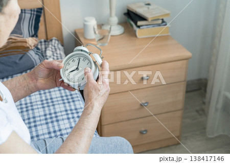 Senior woman is holding an alarm clock in her hands, adjusting time for daily routine, indicating aging and morning lifestyle Senior woman is holding an alarm clock in her hands, adjusting time for daily routine, indicating aging and morning lifestyle 138417146