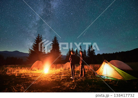 Two hikers stand by campfire under vast, star-filled sky, near tents glowing softly in background. Milky Way stretches overhead, creating magical and serene ambiance in tranquil mountain setting. Two hikers stand by campfire under vast, star-filled sky, near tents glowing softly in background. Milky Way stretches overhead, creating magical and serene ambiance in tranquil mountain setting. 138417732