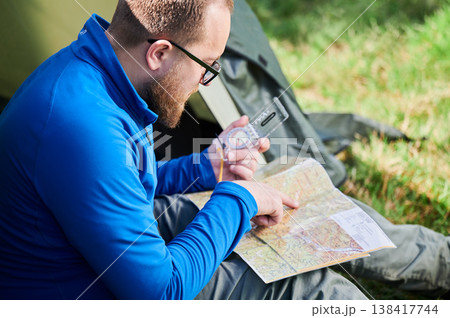 Close up man in glasses closely examines map while holding compass. He sits by his tent, deeply focused on navigation. Lush greenery, suggesting serene outdoor adventure. Close up man in glasses closely examines map while holding compass. He sits by his tent, deeply focused on navigation. Lush greenery, suggesting serene outdoor adventure. 138417744