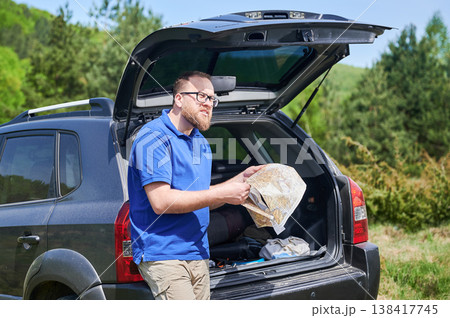 Man tourist leans on open trunk of SUV, deeply focused on map. Traveler wears blue shirt and beige pants, standing in lush, green area, carefully planning or navigating his route. Man tourist leans on open trunk of SUV, deeply focused on map. Traveler wears blue shirt and beige pants, standing in lush, green area, carefully planning or navigating his route. 138417745
