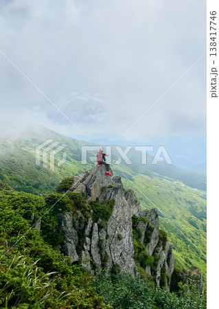 Man hiker in orange jacket and red shoes sits on edge of jagged rock outcrop. Misty, green mountains, cloudy sky create dramatic and awe-inspiring backdrop, highlighting adventurous spirit of scene. Man hiker in orange jacket and red shoes sits on edge of jagged rock outcrop. Misty, green mountains, cloudy sky create dramatic and awe-inspiring backdrop, highlighting adventurous spirit of scene. 138417746